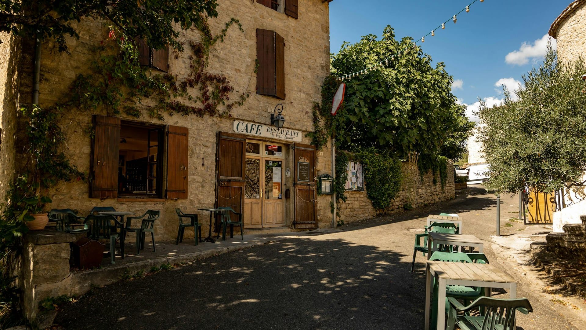 Un café en pierre pittoresque en Provence avec des volets en bois, des tables extérieures et de la verdure dans une rue calme sous un ciel bleu clair.