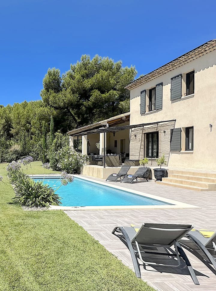 Terrasse d’une villa provençale avec une piscine turquoise, transats modernes et jardin verdoyant, entourée de pins sous un ciel bleu éclatant.