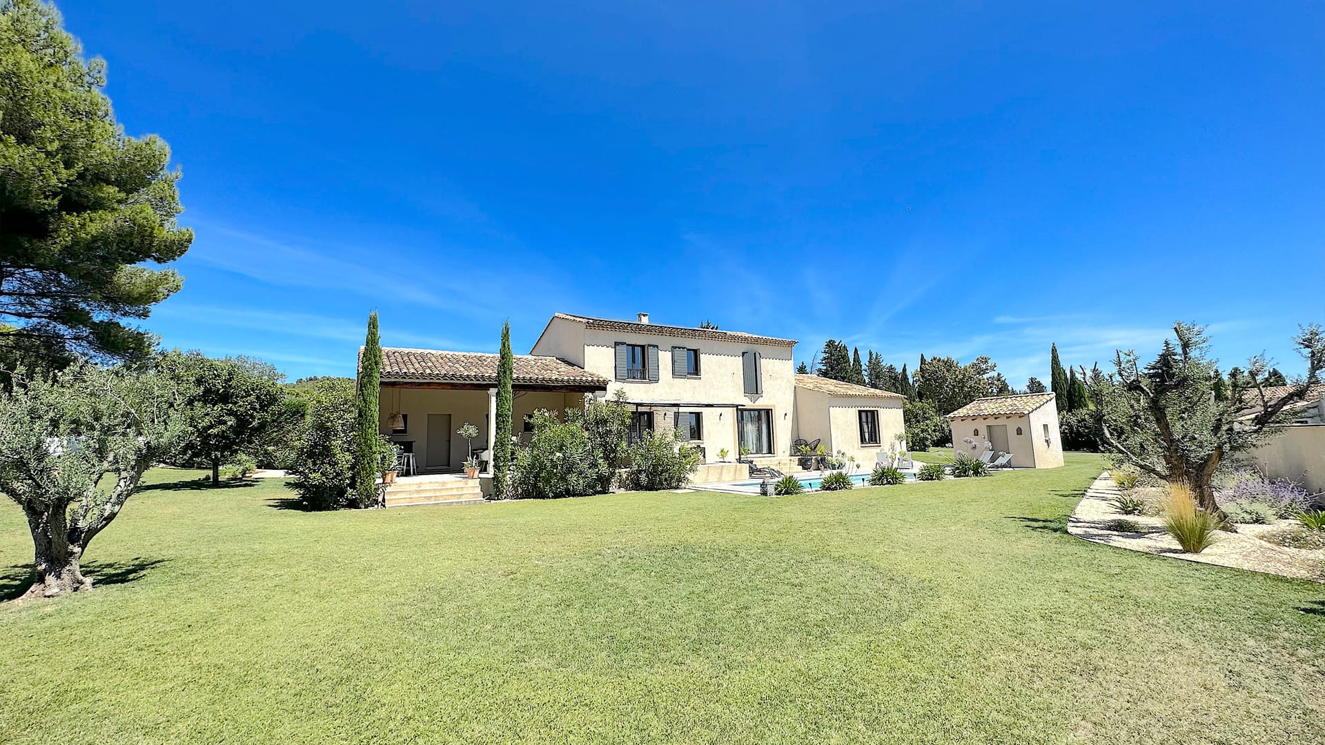 Grande maison provençale avec piscine, pelouse parfaitement entretenue et oliviers, entourée d'un ciel bleu clair.