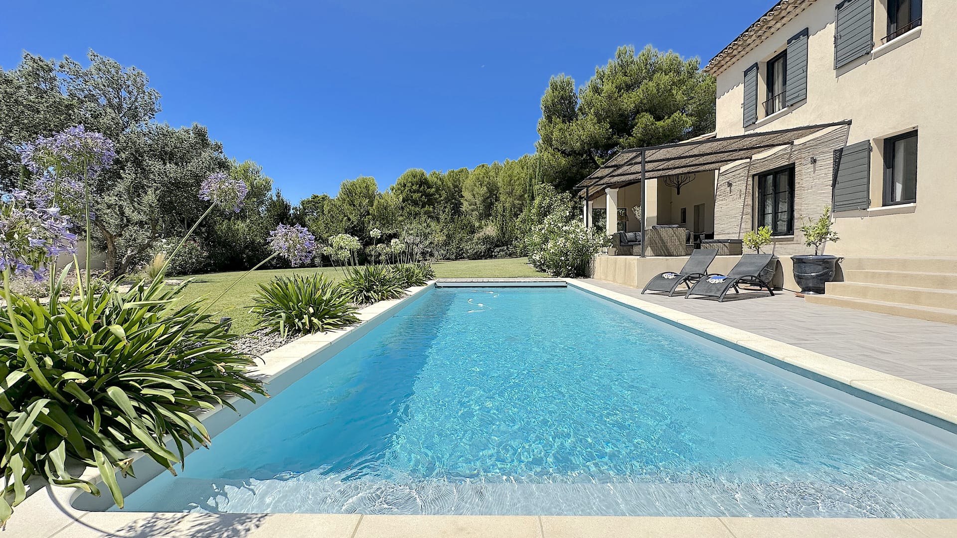 Une piscine extérieure rectangulaire à côté d'une maison beige aux volets verts, entourée d'herbe, d'arbres et de plantes à fleurs sous un ciel bleu clair.