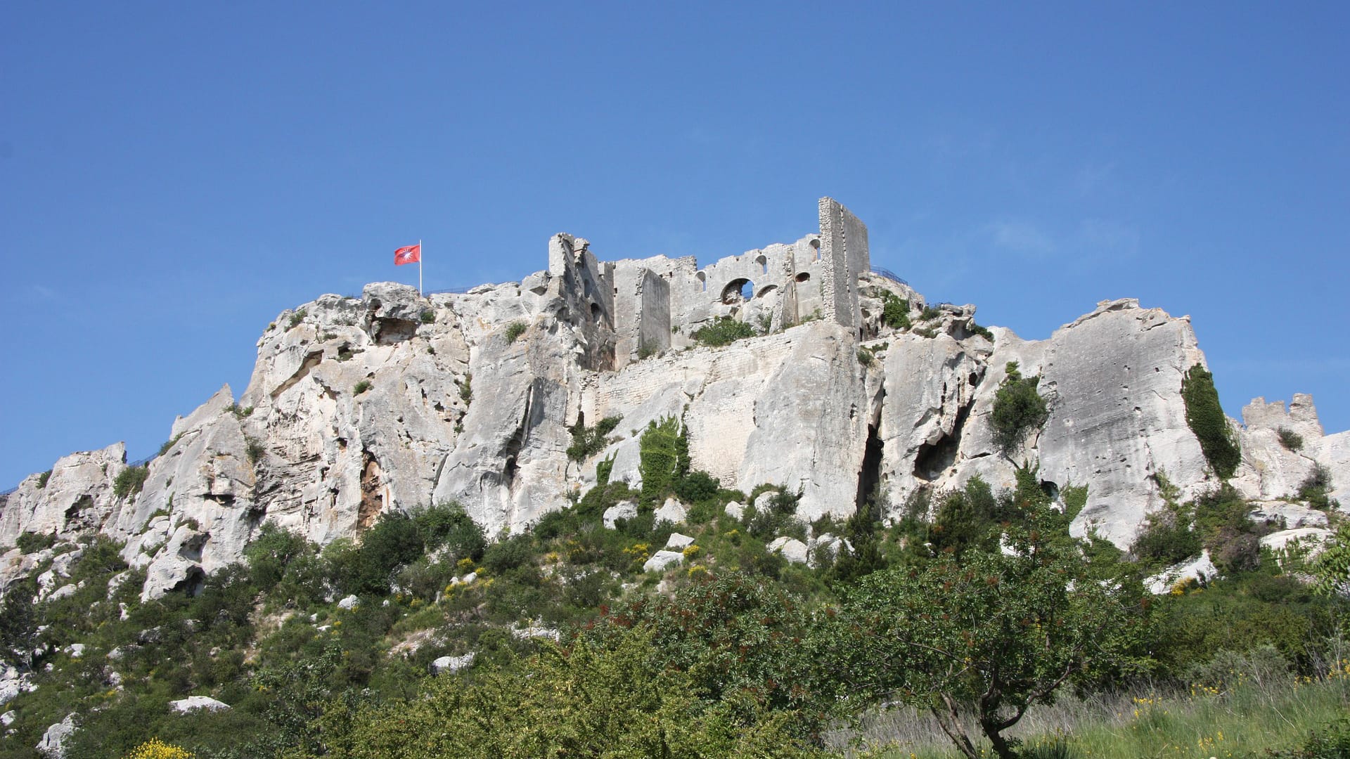 Ruines d’un château médiéval sur une colline rocheuse avec un drapeau rouge flottant sous un ciel bleu clair.