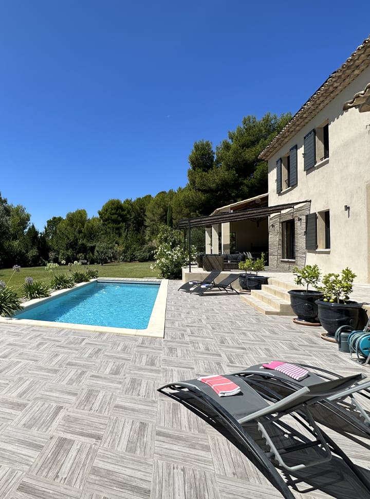 Une maison moderne avec un patio carrelé, des chaises longues, une piscine et un paysage verdoyant sous un ciel bleu clair.