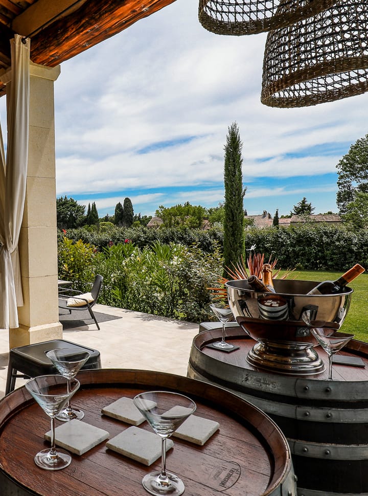 Scène de patio extérieur avec des verres à vin sur des tonneaux, un grand seau à glace et une vue sur un jardin luxuriant sous un ciel partiellement nuageux.